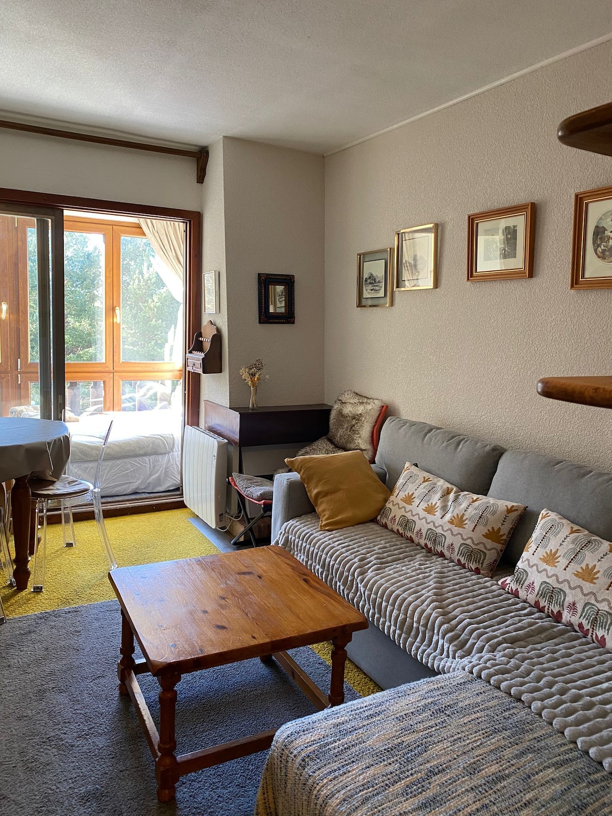 A comfortable living area features a gray sofa adorned with patterned cushions. A wooden table is positioned in front, complemented by a warm yellow carpet. Sunlight filters through large glass doors leading to a balcony, enhancing the inviting atmosphere.