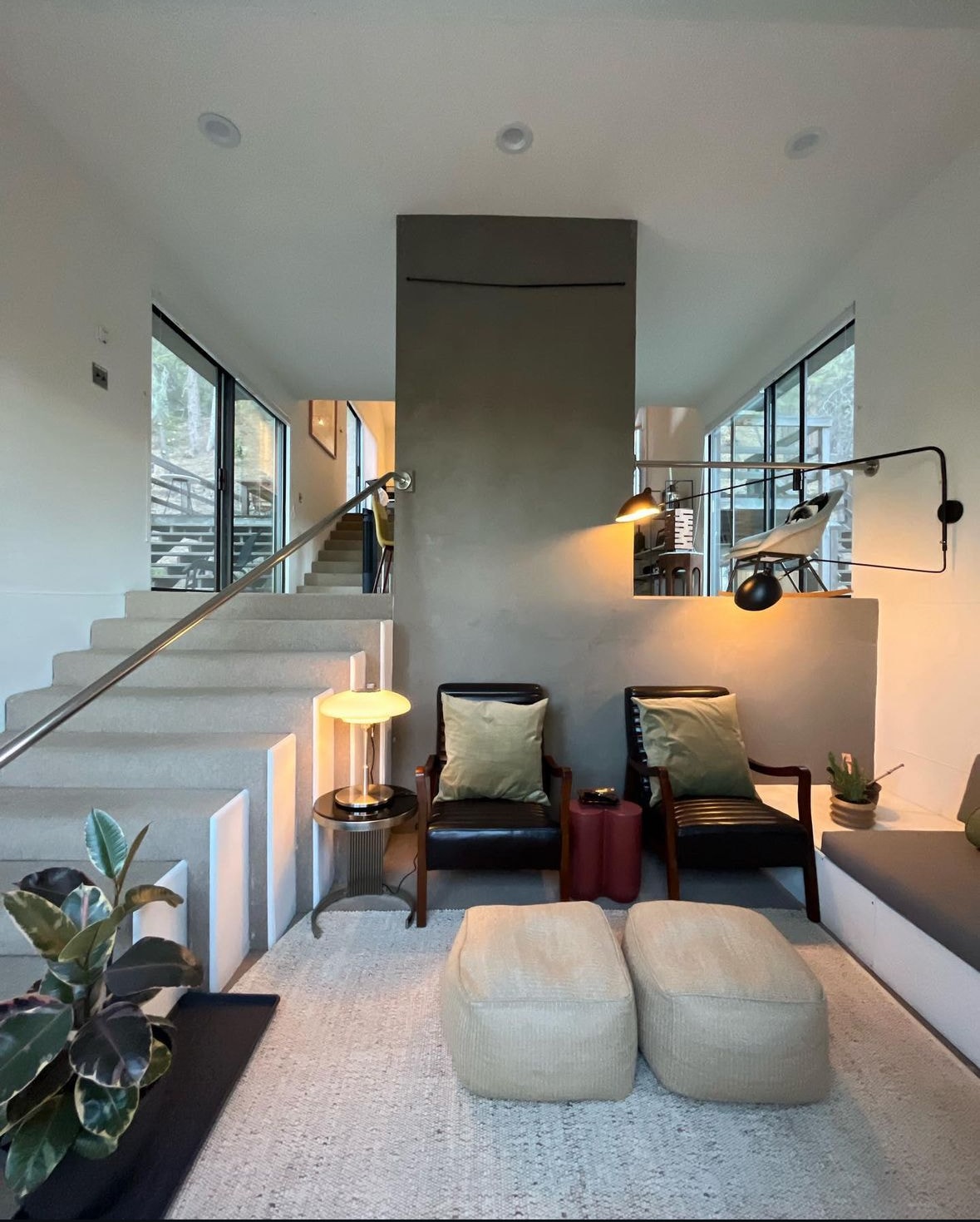 A modern living area is showcased, featuring a staircase leading to the upper level. Two sleek black armchairs are situated beside a small side table with a lamp. A potted plant and decorative cushions add natural elements near a cozy seating nook.