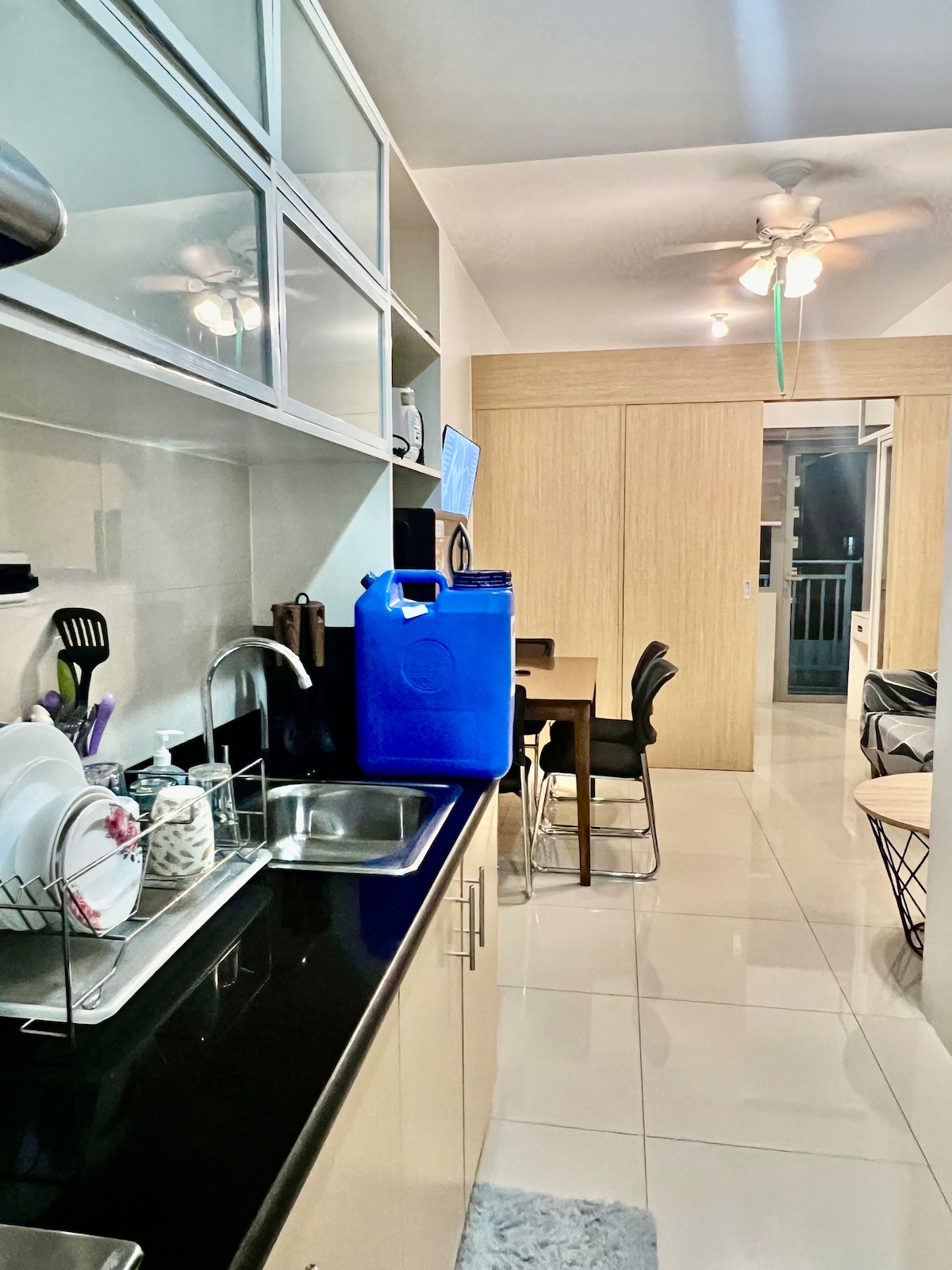 A modern kitchen area features sleek cabinets and a black countertop. Cooking utensils and dishware are neatly arranged. A dining table with four chairs is visible, alongside a window that invites natural light. A ceiling fan is positioned above, enhancing airflow.
