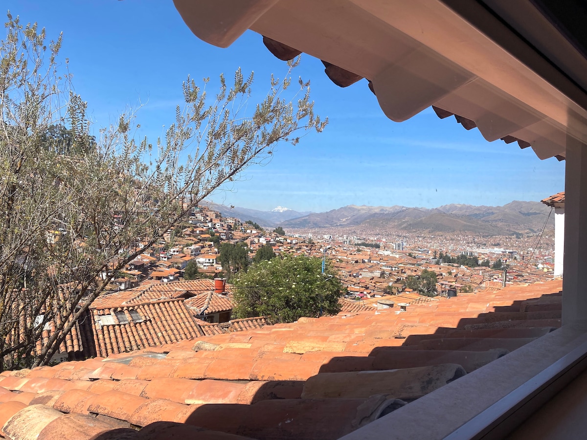A scenic view of Cusco is framed by a window, highlighting the city’s terracotta roofs and distant mountains. The landscape is dotted with green trees, showcasing the natural beauty surrounding the urban area under a clear blue sky.