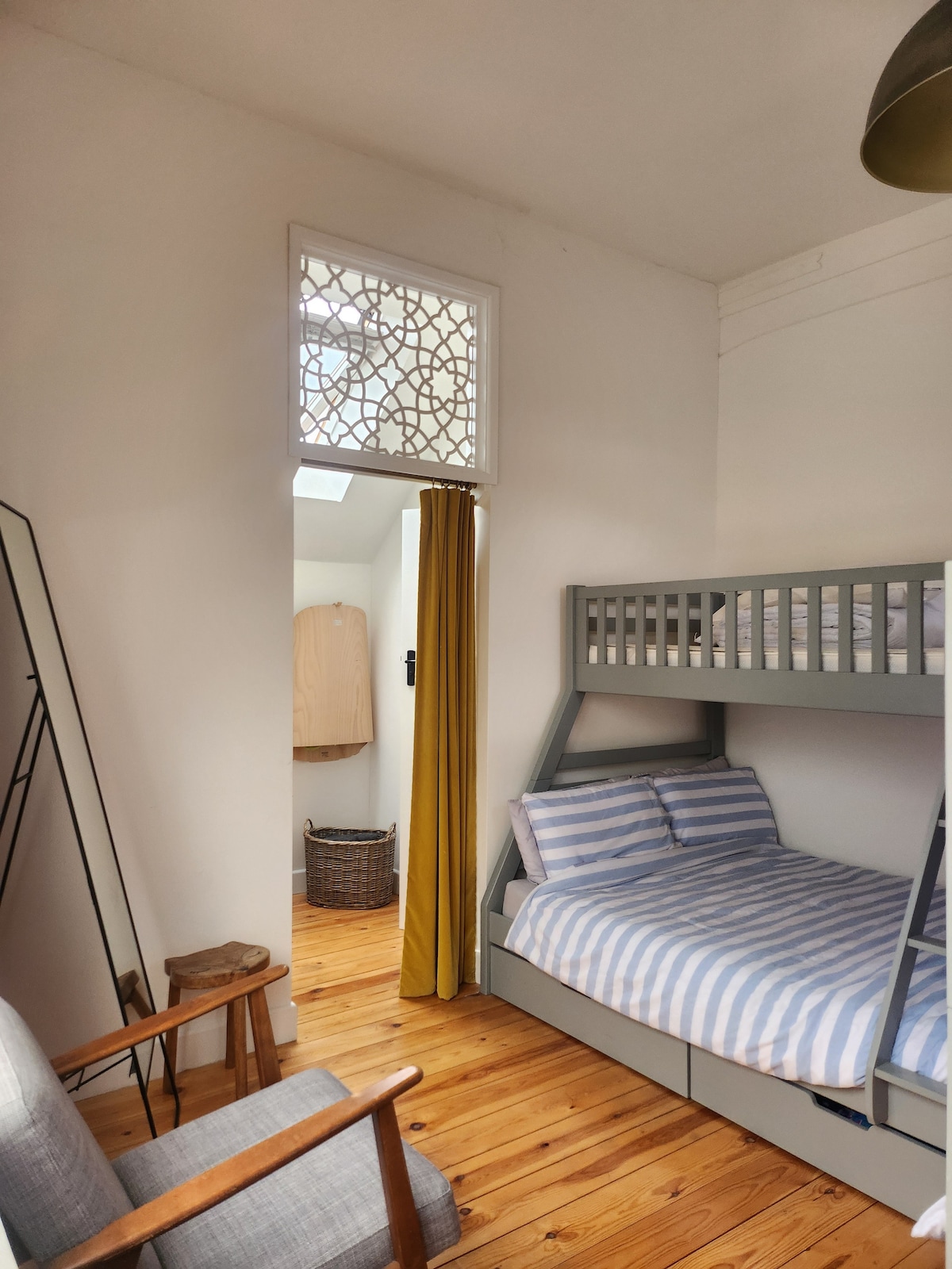 A cozy sleeping area features a double bunk with striped bedding and a circular window illuminating the space. A small chair and side table are positioned nearby, enhancing the room's functional layout. Light wood flooring adds warmth to the atmosphere.