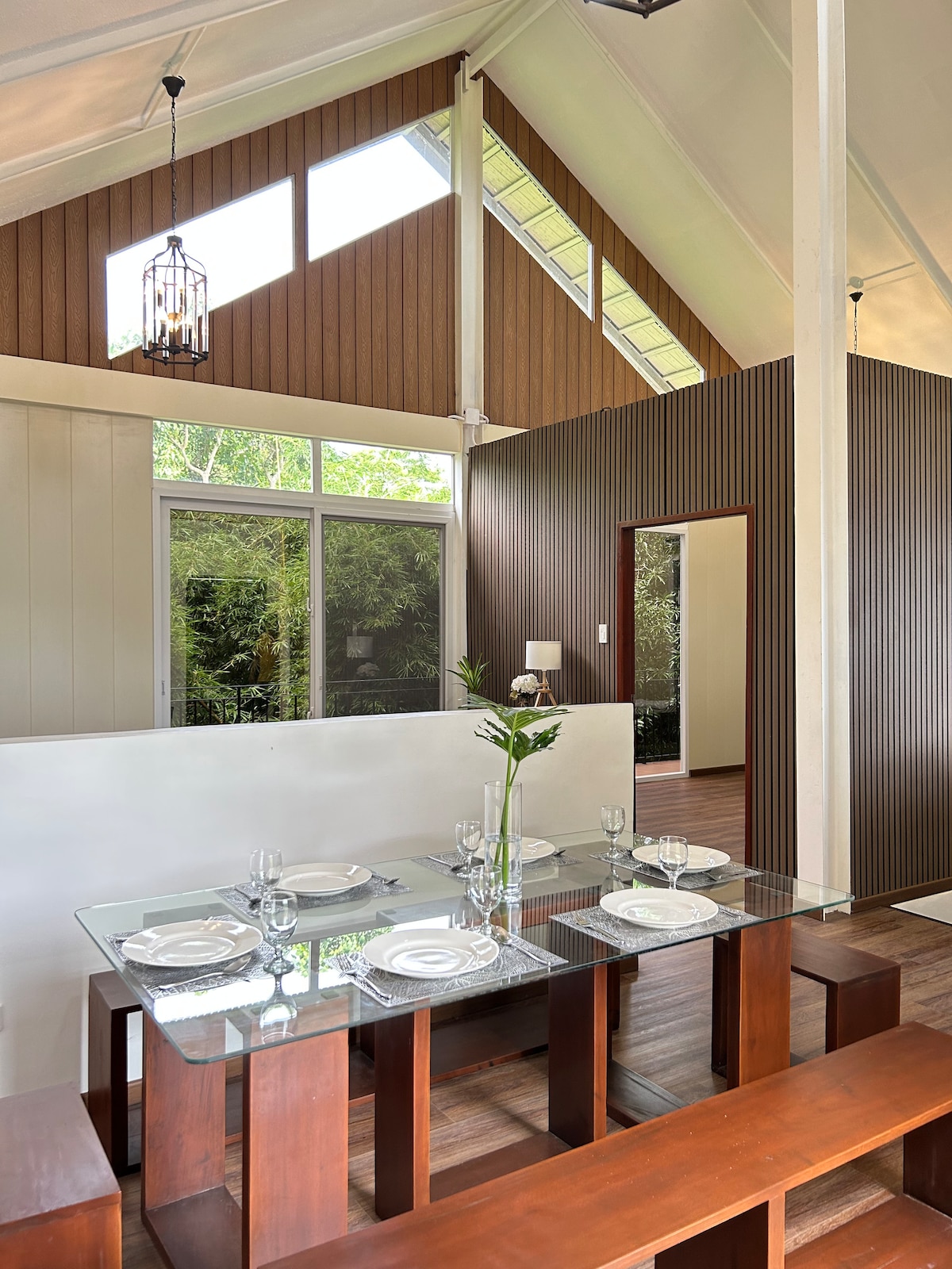 A modern dining area features a glass-topped table set for four. Elegant dishware and glassware are arranged neatly. High ceilings are complemented by large windows allowing natural light to fill the space, while wood accents add warmth to the contemporary design.