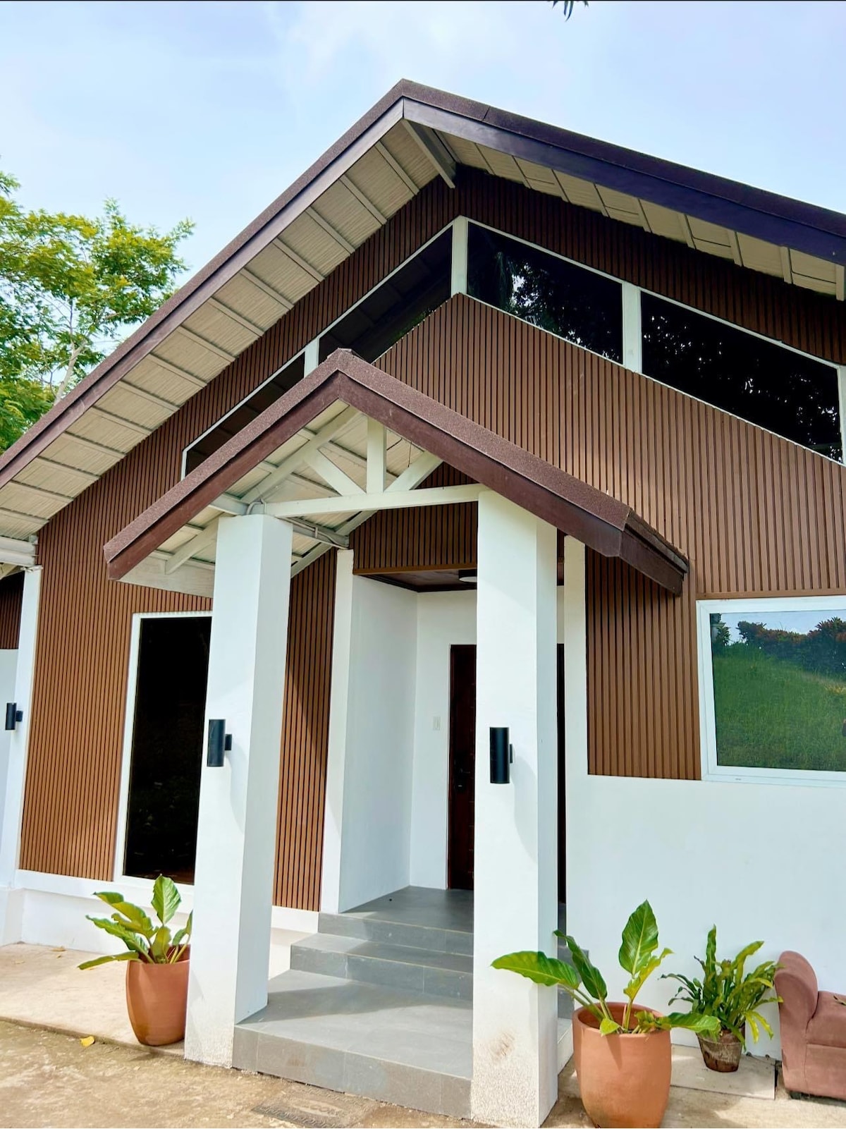 The entrance of the newly built villa features a sloped roof and a covered porch. Two potted plants are placed on either side of the entrance. Large windows allow natural light to illuminate the space, complemented by the earthy tones of the exterior.