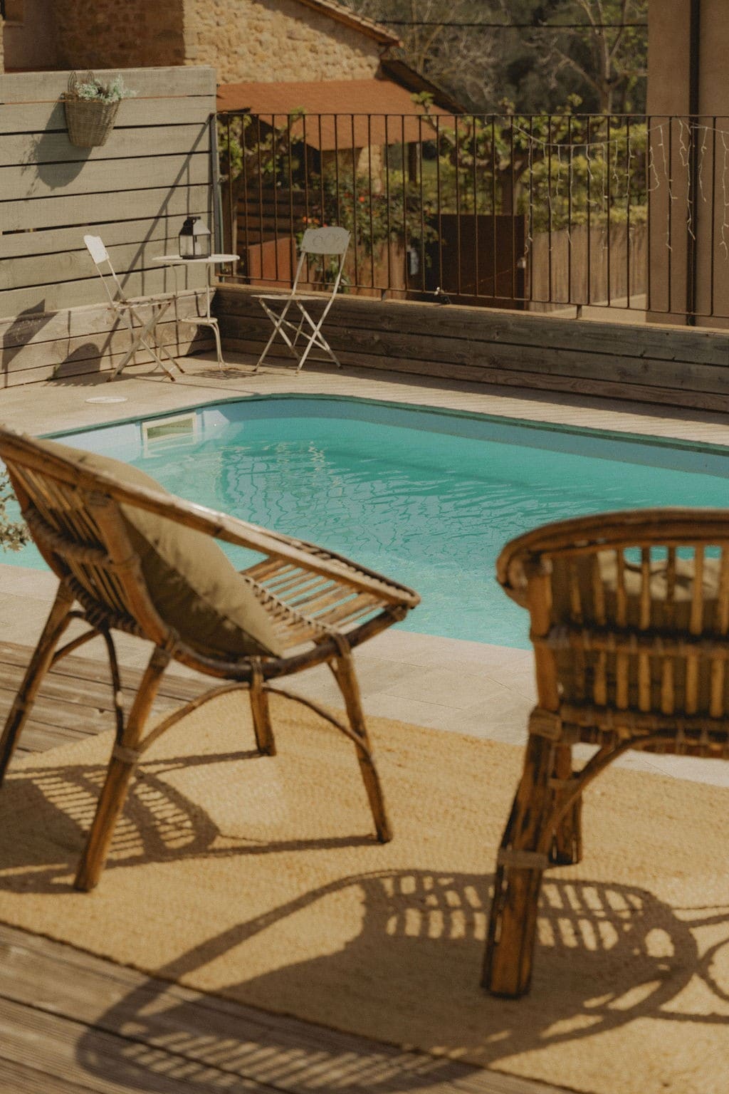 An inviting pool area features a rectangular swimming pool surrounded by wooden decking. Two wicker chairs with cushions are positioned nearby on a soft rug. The setting is complemented by a low railing and partial views of surrounding greenery.