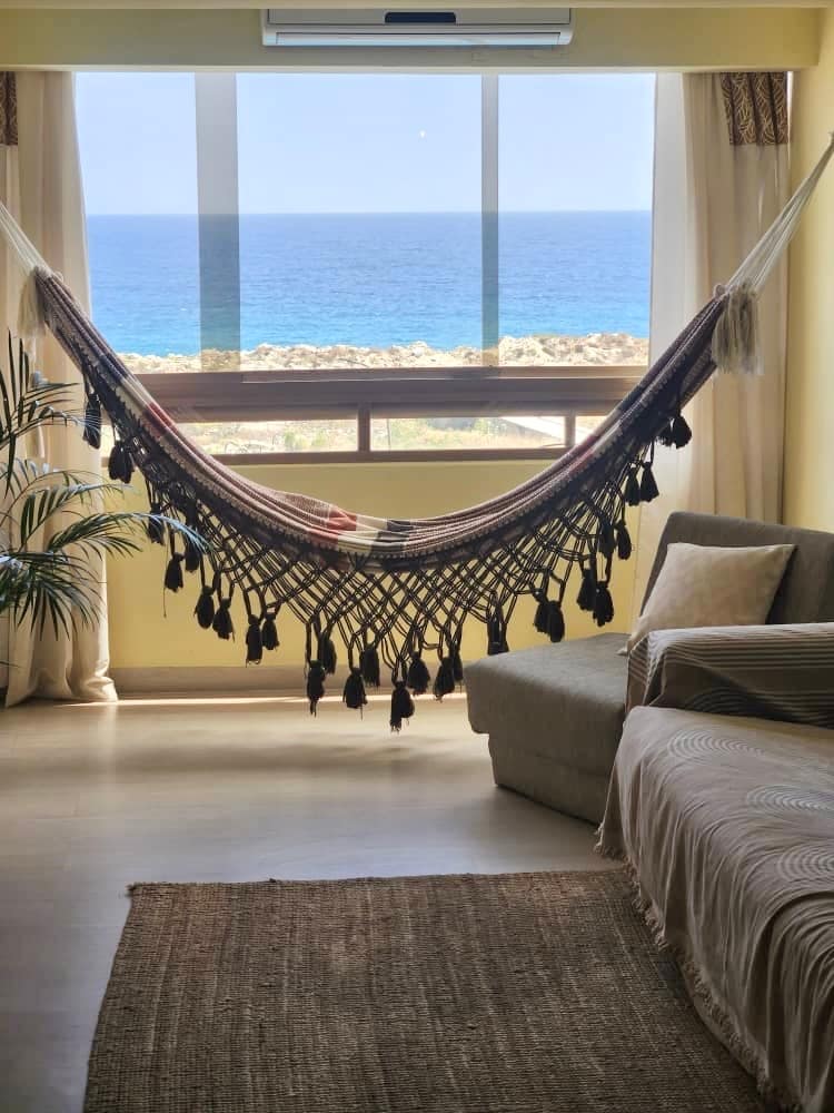 A relaxing living area features a spacious sofa and a light-colored rug. A hammock is suspended near a large window, providing a view of the sea. Natural light fills the room, accentuated by the soft curtains that frame the window.