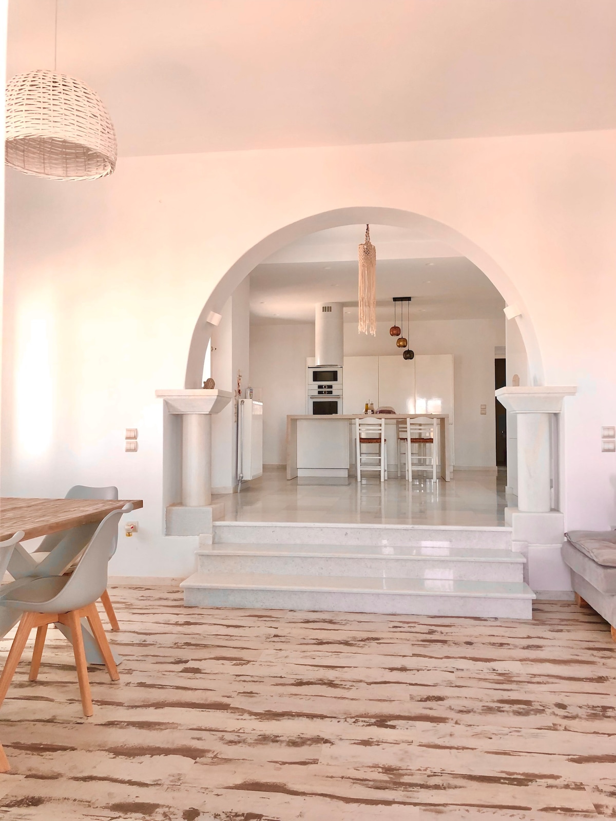 An open-concept living area features a clear view of the modern kitchen behind an elegant arch. A wooden dining table is surrounded by light-colored chairs, while a cozy seating area with a sofa can be seen to the right. Natural light enhances the bright ambiance.