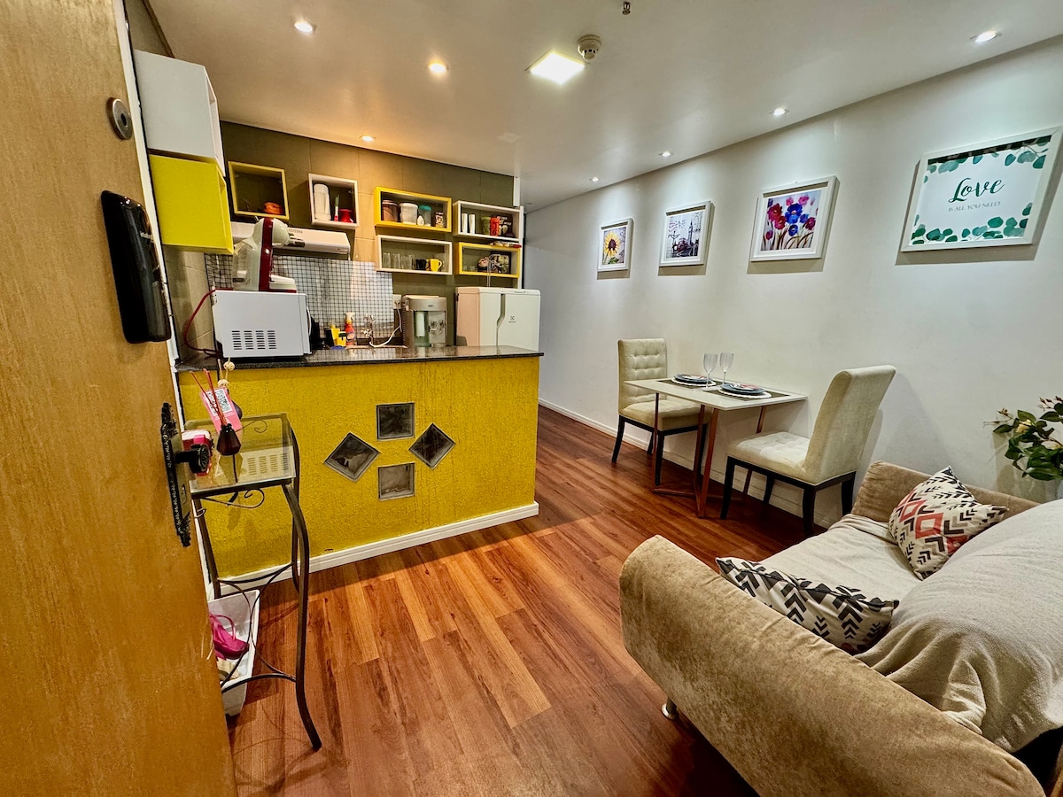 The open living space features a compact kitchen equipped with essential appliances, including a microwave and coffee maker. A small dining table for two is located next to a cozy seating area adorned with decorative pillows. Bright artwork complements the neutral walls, creating a vibrant atmosphere.