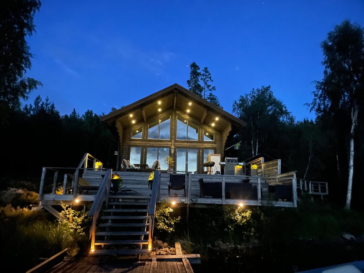 The lodge is illuminated at night, showcasing its wooden exterior and expansive glass windows. A staircase leads to a spacious deck area, featuring outdoor seating and plants. Surrounding trees create a serene backdrop, enhancing the peaceful atmosphere of this lakeside retreat.