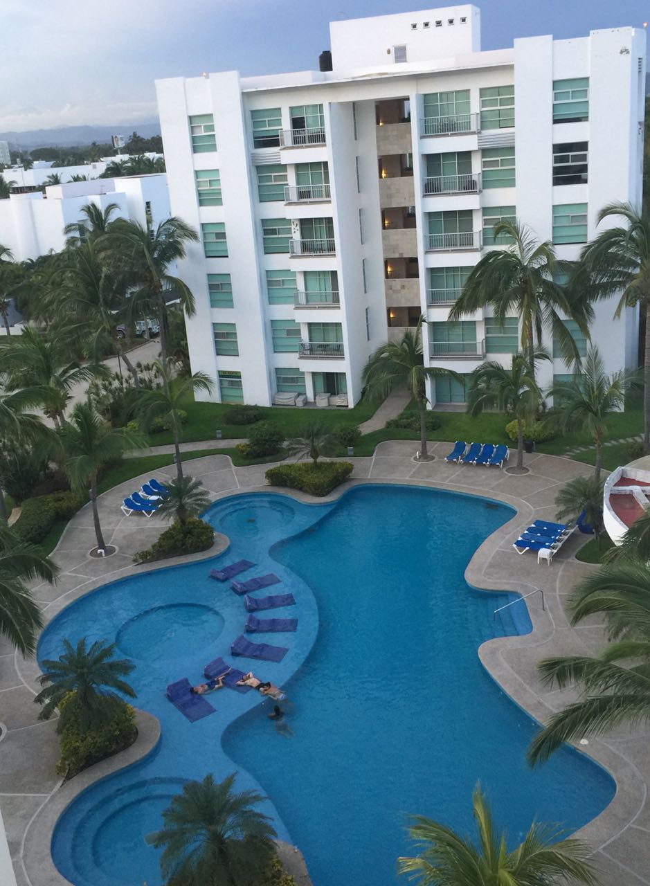 An aerial view showcases a large, blue swimming pool integrated with a zigzag design, surrounded by tropical palm trees. Lounge chairs are positioned around the pool area, offering a relaxing outdoor space. The adjacent white building features multiple glass balconies, enhancing the tranquil setting.
