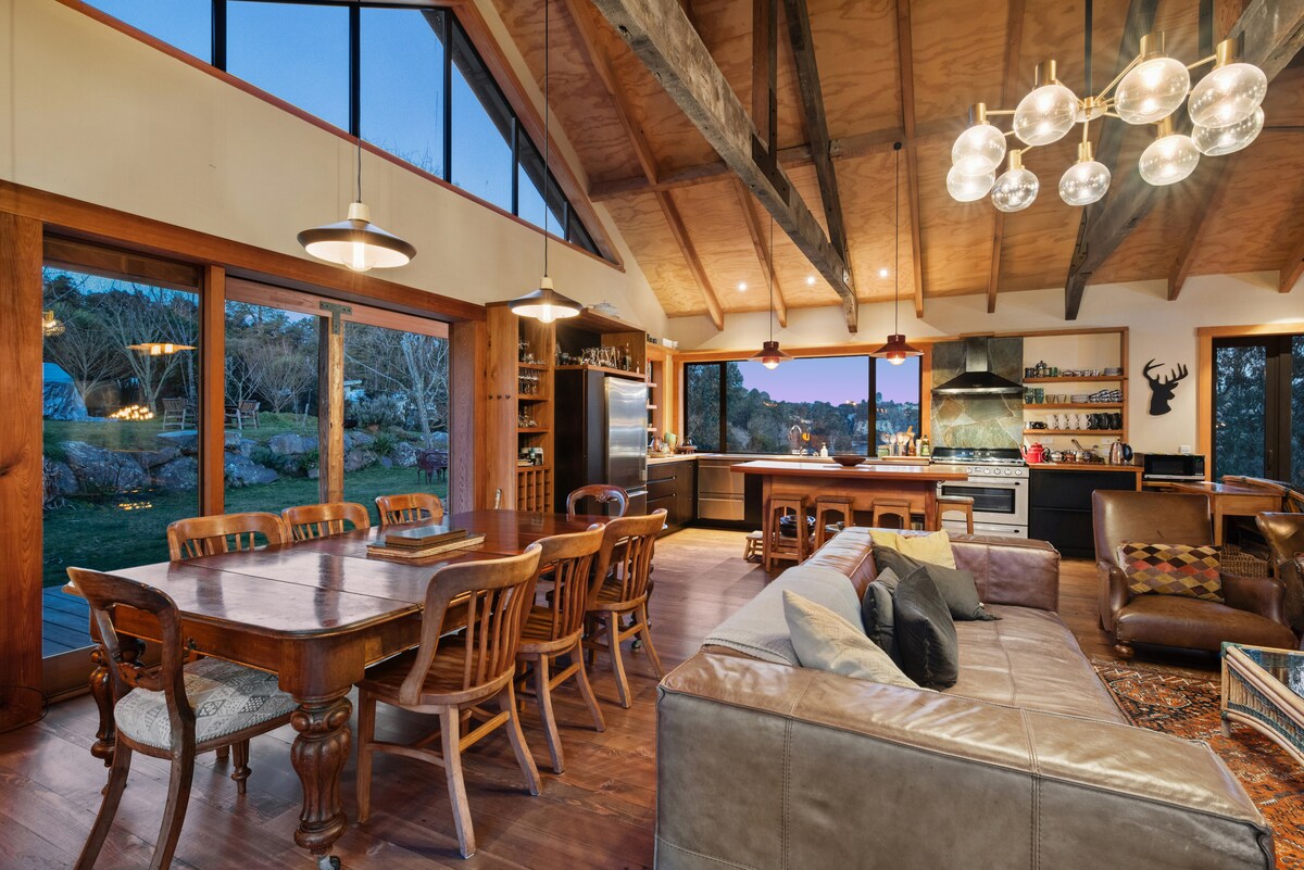 An open-plan living area is showcased with wooden beams and large windows allowing for abundant natural light. A spacious dining table sits surrounded by wooden chairs, while a well-equipped kitchen and inviting seating area can be seen nearby, creating a warm and communal atmosphere.