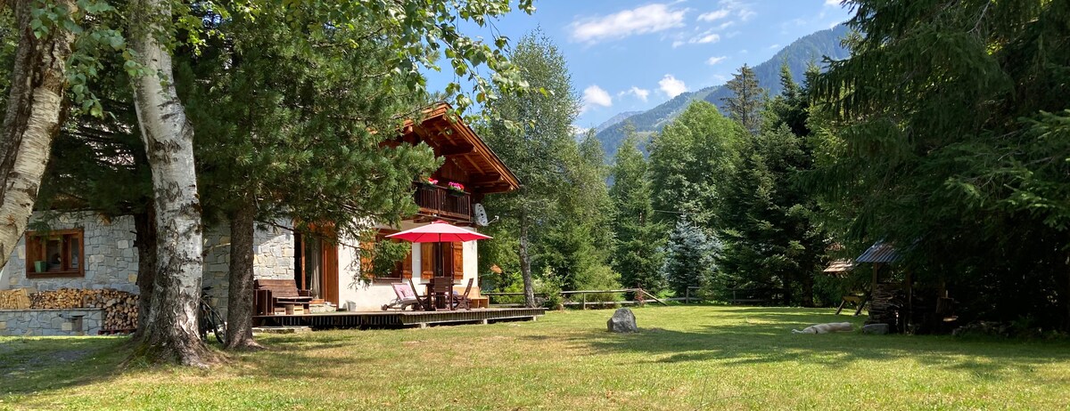 The exterior of the chalet is surrounded by lush greenery and features a wooden terrace with a red umbrella providing shaded seating. Nearby, a grassy area can be seen with trees framing the scene and mountains in the background, offering a peaceful and natural setting.