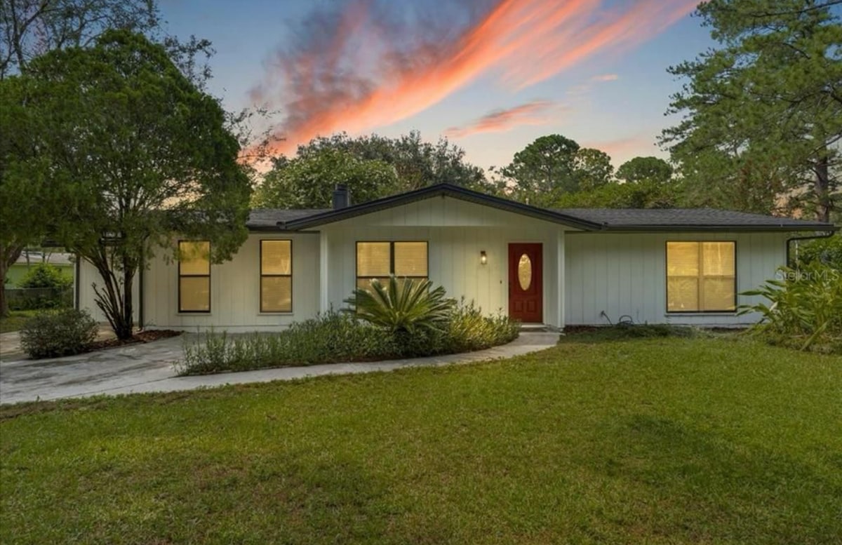 A single-story home is set against a colorful sunset. The light blue exterior is complemented by large windows, showcasing the surrounding greenery. A pathway leads to a welcoming front door, while a well-maintained lawn adds to the home's inviting presence.