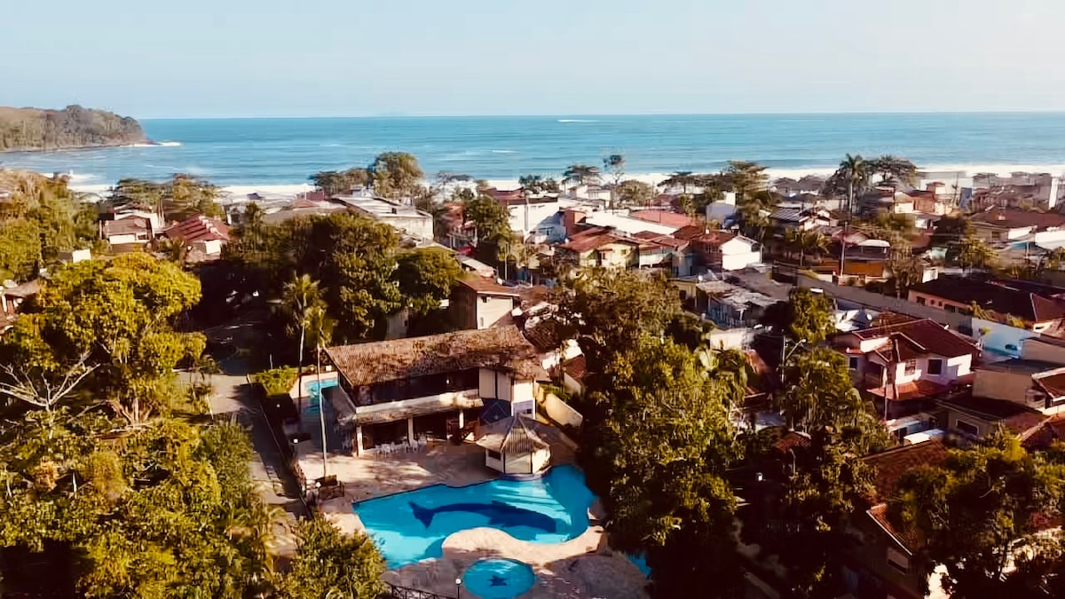 An aerial view captures the vibrant community of Maresias, with lush greenery surrounding a blue pool area. The ocean is visible in the background, and the colorful rooftops of nearby houses create a lively coastal atmosphere.