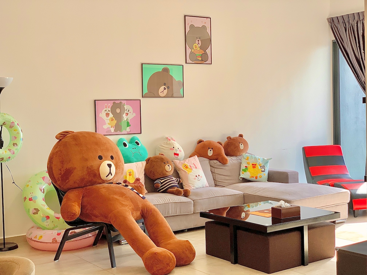 A cheerful living room features a plush bear character sitting on a chair beside a modern sofa. Colorful cushions and framed artwork depicting friendly characters adorn the space. The large windows allow natural light to fill the area, enhancing the inviting ambiance.