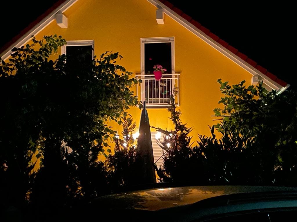 The exterior of a yellow house is illuminated at night, showcasing a balcony adorned with a flowering pot. Surrounding greenery adds to the serene ambiance, while soft lighting enhances the inviting appearance of the structure.