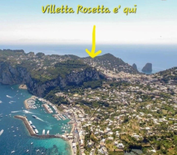 An aerial view highlights the picturesque coastline of Capri, featuring the marina, lush greenery, and the iconic Faraglioni rocks in the distance. The image is marked to indicate the location of Villetta Rosetta, showcasing its positioning within the beautiful landscape.