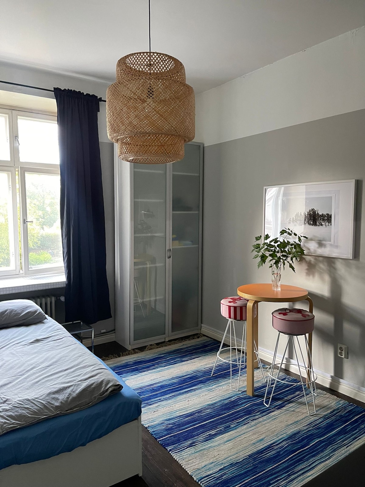 A single room features a spacious bed with a light blue blanket, a stylish woven pendant light, and a small round table with a plant. Two stools with vibrant seats are positioned beside the table. A glass-fronted cabinet and a large window with dark curtains are also visible.