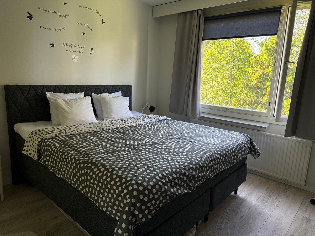 A cozy bedroom includes a spacious bed adorned with a dotted blanket and crisp white pillows. Natural light enters through a large window with dark blinds, while soft gray curtains frame the view of green trees outside.