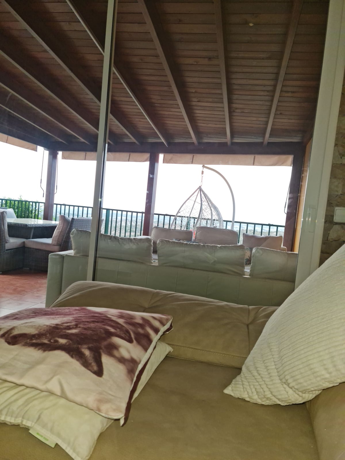 A covered terrace area is visible, featuring a hanging chair and comfortable seating arranged around a table. The wooden ceiling adds warmth, and large windows allow natural light to flood the space while providing a view of the surrounding landscape.