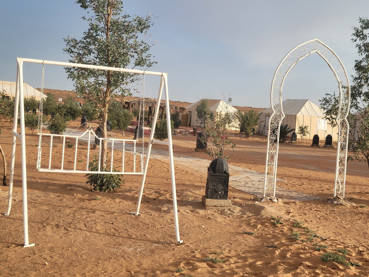 A white swing set sits on sandy ground, framed by greenery. In the background, spacious tents are visible, surrounded by desert flora. An ornate archway leads to a pathway, creating an inviting entrance to the seating area of the camp.