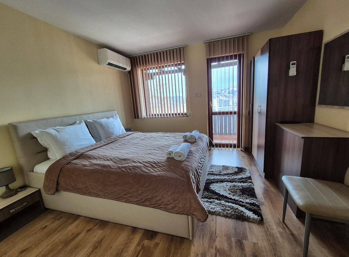 A spacious bedroom features a large bed with neatly arranged towels. Sunlight enters through vertical blinds, casting defined shadows on the wood flooring. A small seating area and a mirrored dresser add functionality, while a balcony door offers views of the surroundings.