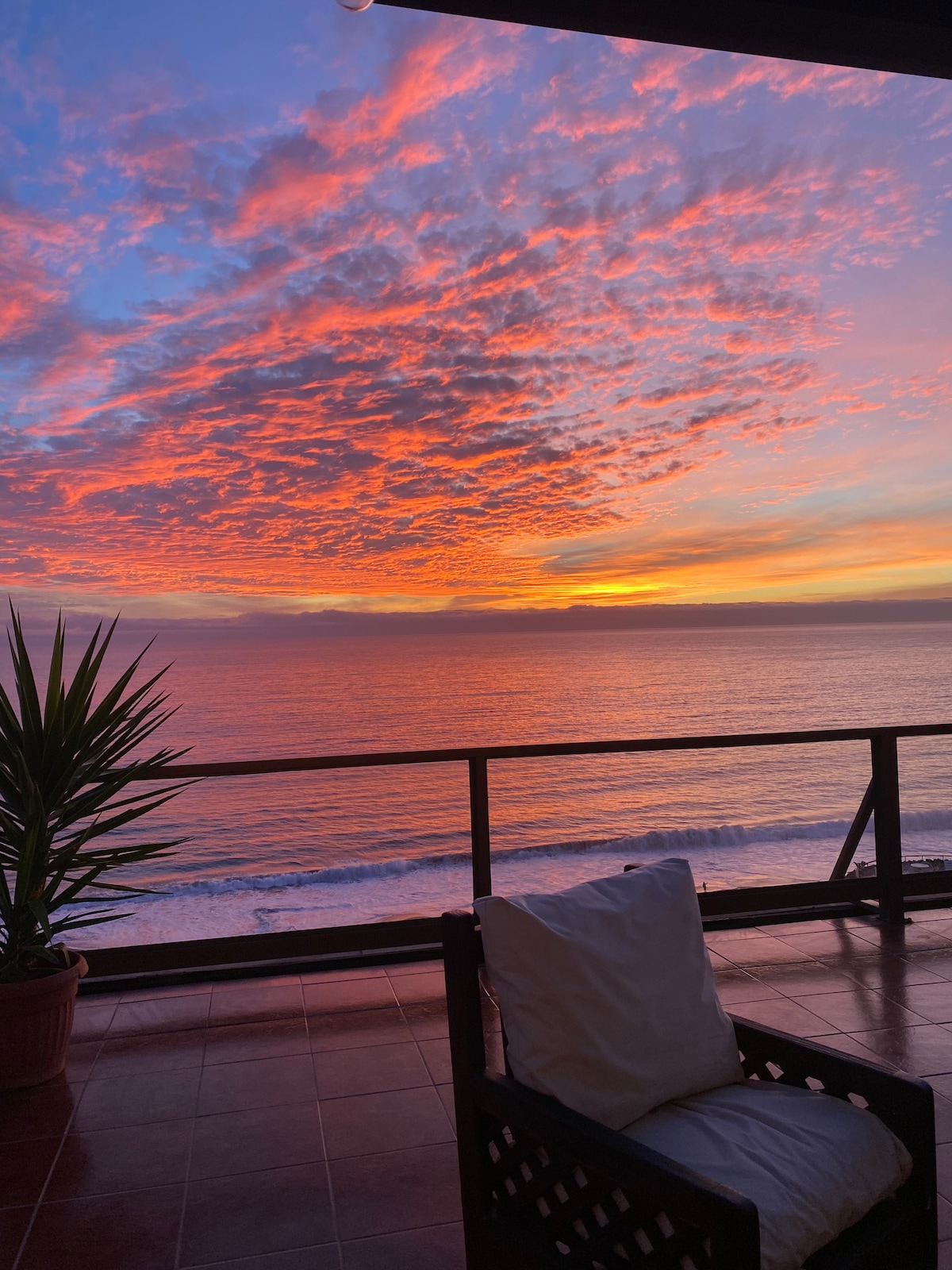 A stunning sunset over the ocean is captured from a spacious terrace. The vibrant hues of orange and pink paint the sky, while gentle waves can be seen in the distance. A solitary chair sits on the terrace, accompanied by a potted plant, enhancing the serene view.