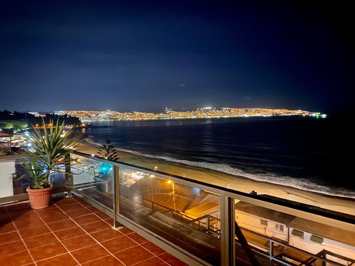 The image features a night view from a large terrace, displaying a panoramic vista of the illuminated coastline and serene ocean waves. A potted plant adds a touch of greenery to the space, while the soft glow of city lights reflects off the water.