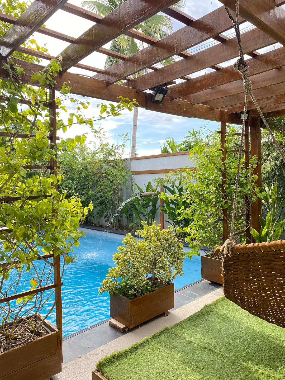 A serene swimming pool is framed by lush greenery, with wooden planters filled with plants enhancing the tranquil setting. A swing chair is positioned nearby, overlooking the water and providing a peaceful spot to relax. Natural light filters through the surrounding foliage.