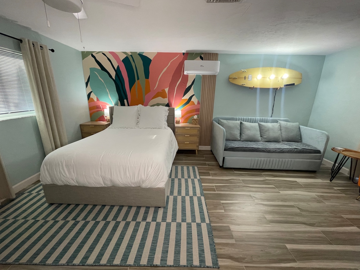 A cozy studio space is presented with a spacious bed flanked by nightstands and reading lamps. A stylish mural in vibrant colors adds visual interest to one wall. A comfortable daybed and a small side table complement the setting, with soft lighting enhancing the relaxed ambiance.