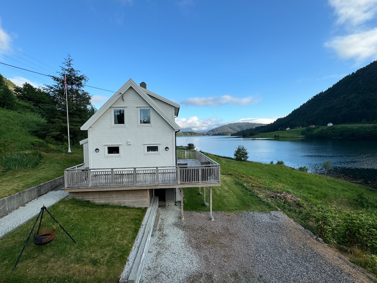 The two-story house is situated by a tranquil fjord, featuring a spacious wooden terrace overlooking the water. Lush greenery surrounds the property, with rolling hills in the background. A gravel parking area is visible, providing access to the scenic outdoor environment.