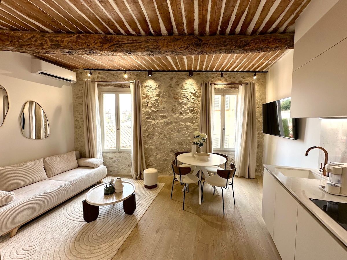 A bright living space features exposed beams and stone walls. A comfortable sofa is positioned alongside a circular dining table. Natural light pours in through two windows, while decorative mirrors add visual interest to the room. The area is accentuated by light-colored flooring.