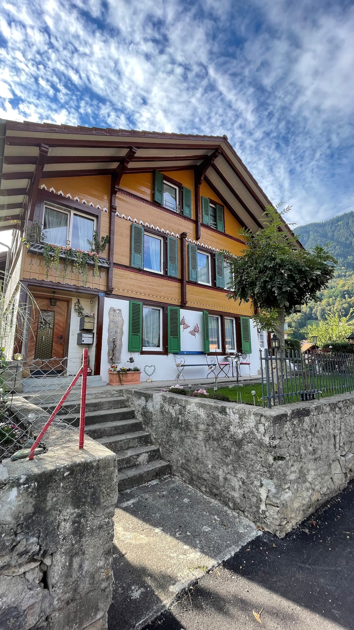 A charming chalet-style building features distinct yellow and brown wood siding, accented by green shutters. A well-maintained front yard includes a small tree and a pathway leading up to the entrance. The surrounding landscape showcases a picturesque mountainous backdrop under a partly cloudy sky.