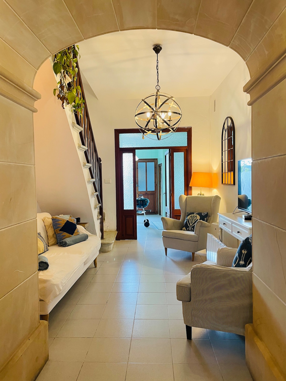 An arched entrance leads into a bright hallway featuring a light fixture and comfortable seating options. A staircase is visible on the left side, while a cozy sofa and a TV stand are positioned along the wall. Natural light fills the space through glass doors at the end.
