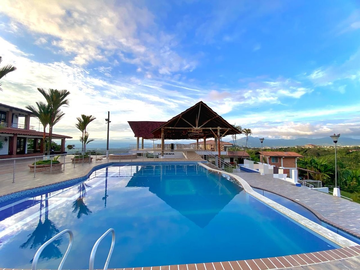 A spacious swimming pool reflects the sky's colors, bordered by a sunlit patio. A large, open pavilion stands nearby, offering shaded seating areas. Palm trees add greenery to the scene, while distant hills create a scenic backdrop.