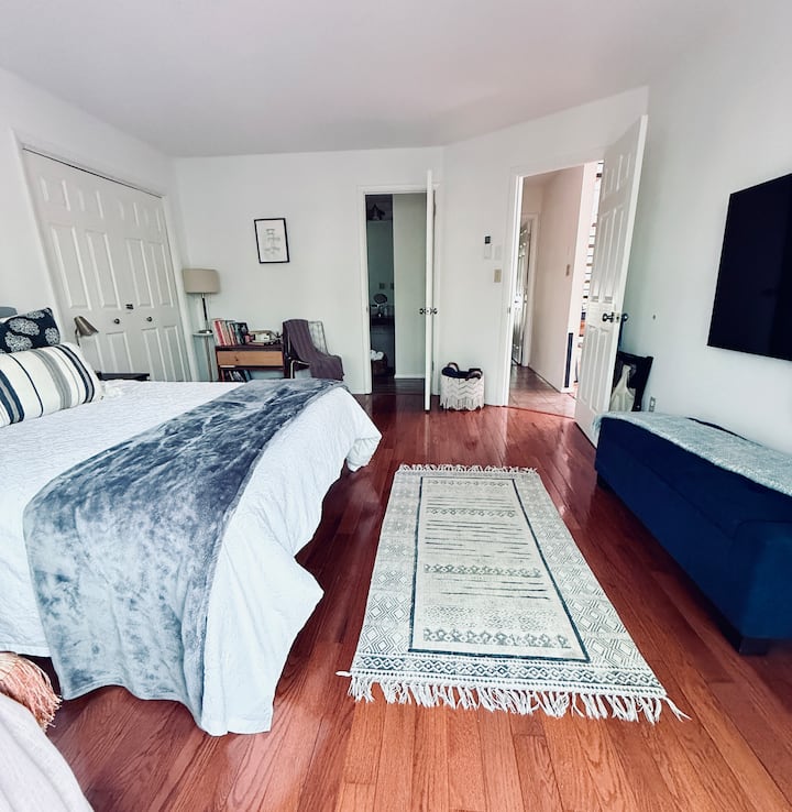 Sunlit suite featuring warm wood floors and sliding glass doors that open to a leafy balcony. A sleek upholstered bench, wall-mounted TV, and understated décor offer a peaceful, modern haven for relaxation.