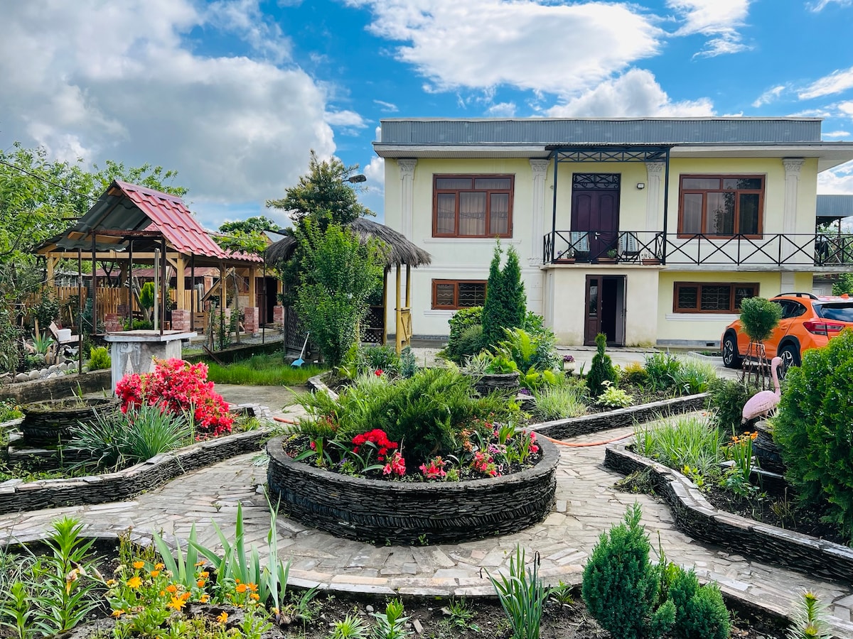 The exterior showcases a well-maintained garden with various plants and vibrant flowers, framed by stone pathways. The two-story house features a spacious porch with a decorative railing and large windows. A thatched gazebo and patio with seating can also be seen in the background.
