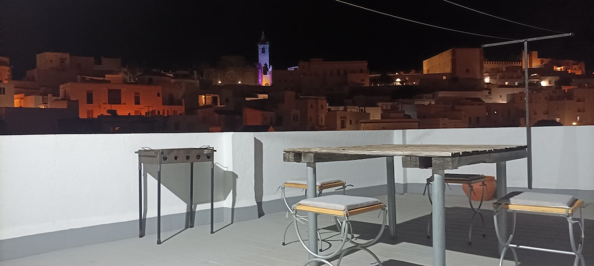 A rooftop terrace is featured under a night sky, showcasing distant illuminated buildings with a tower standing prominently. A wooden table with chairs is arranged for outdoor dining, along with a small grill, providing a spacious area for relaxation and enjoying the night views.