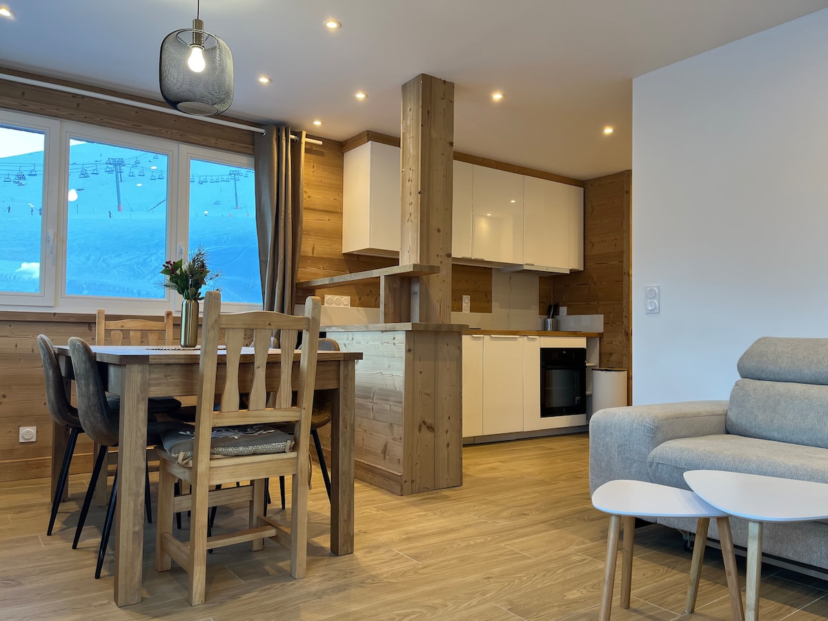 The open-plan living area features a light wooden dining table surrounded by chairs, positioned near a window with scenic views. A modern kitchen is equipped with essential appliances. Comfortable seating is arranged in the living space, complemented by natural light streaming through the large windows.