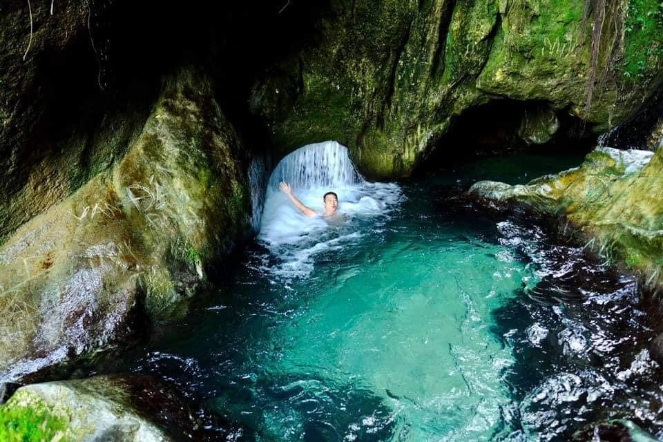 A person is enjoying a tranquil moment in a natural swimming area surrounded by smooth, moss-covered rocks. Clear water flows gently over the stones, creating a serene atmosphere. Lush greenery frames the scene, enhancing the connection to nature.