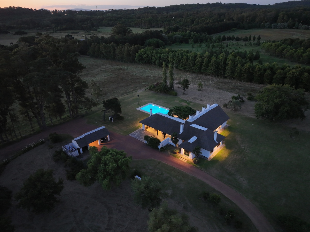 An aerial view captures the expansive property featuring a large house surrounded by open fields and lush trees. A swimming pool glows softly in the evening light, emphasizing the tranquility of the setting.