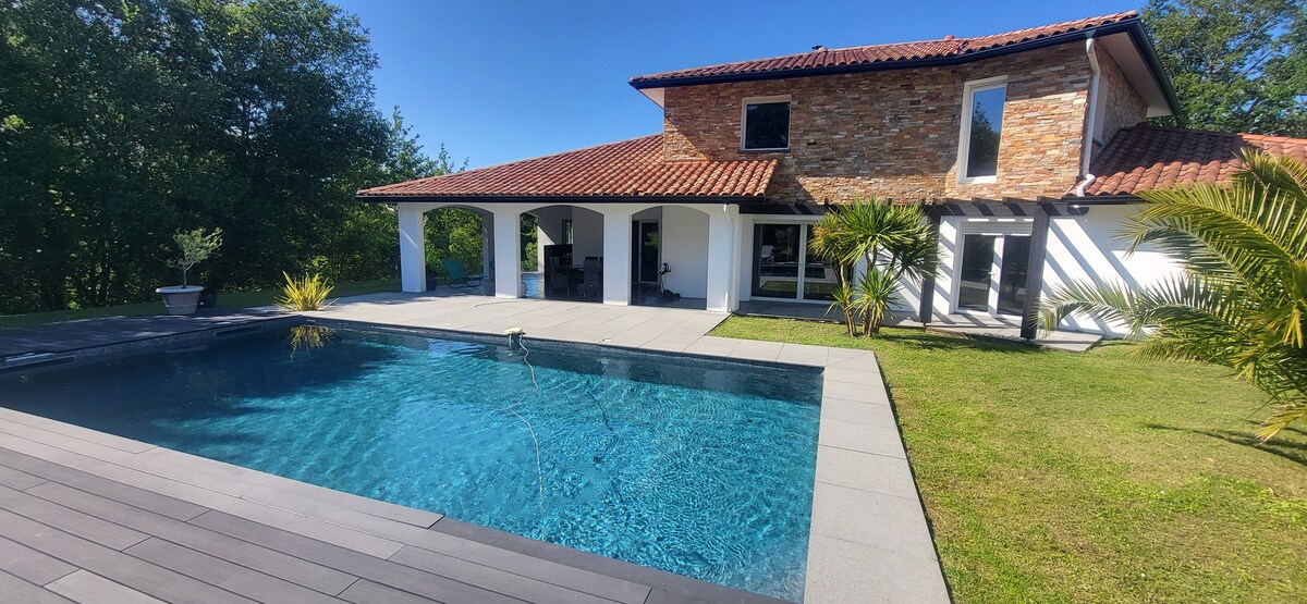 The exterior of the villa features a spacious swimming pool with clear blue water, surrounded by paving stones and a grassy area. The structure showcases a combination of stone and white walls, complemented by a tiled roof and large windows, set against a backdrop of lush greenery.