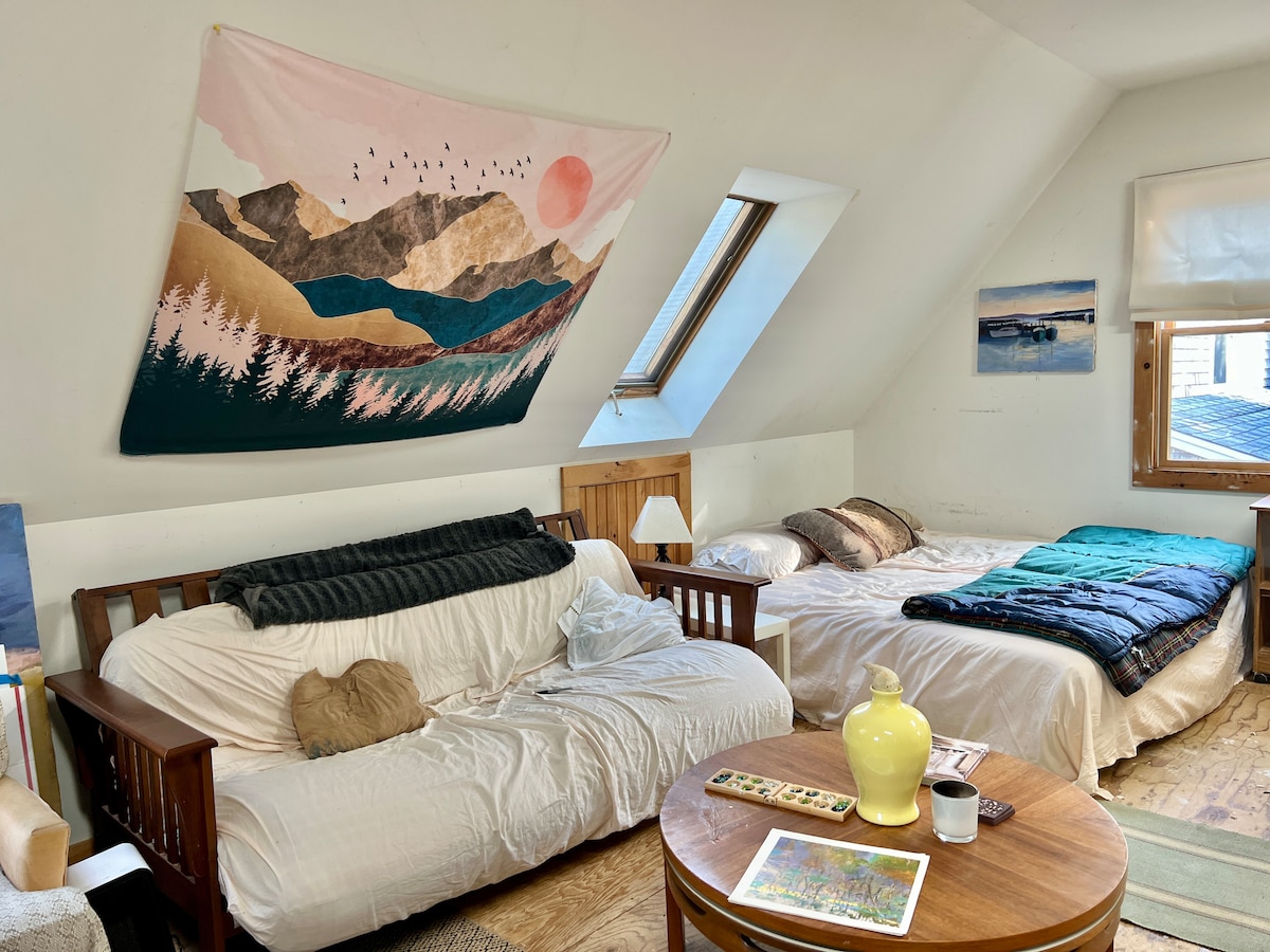 A spacious studio features a combination of a full-size futon and a king bed, layered with neutral bedding. Large windows provide natural light, while a decorative wall hanging adds a touch of character. A round table sits in the center, accompanied by a bright decorative lamp.