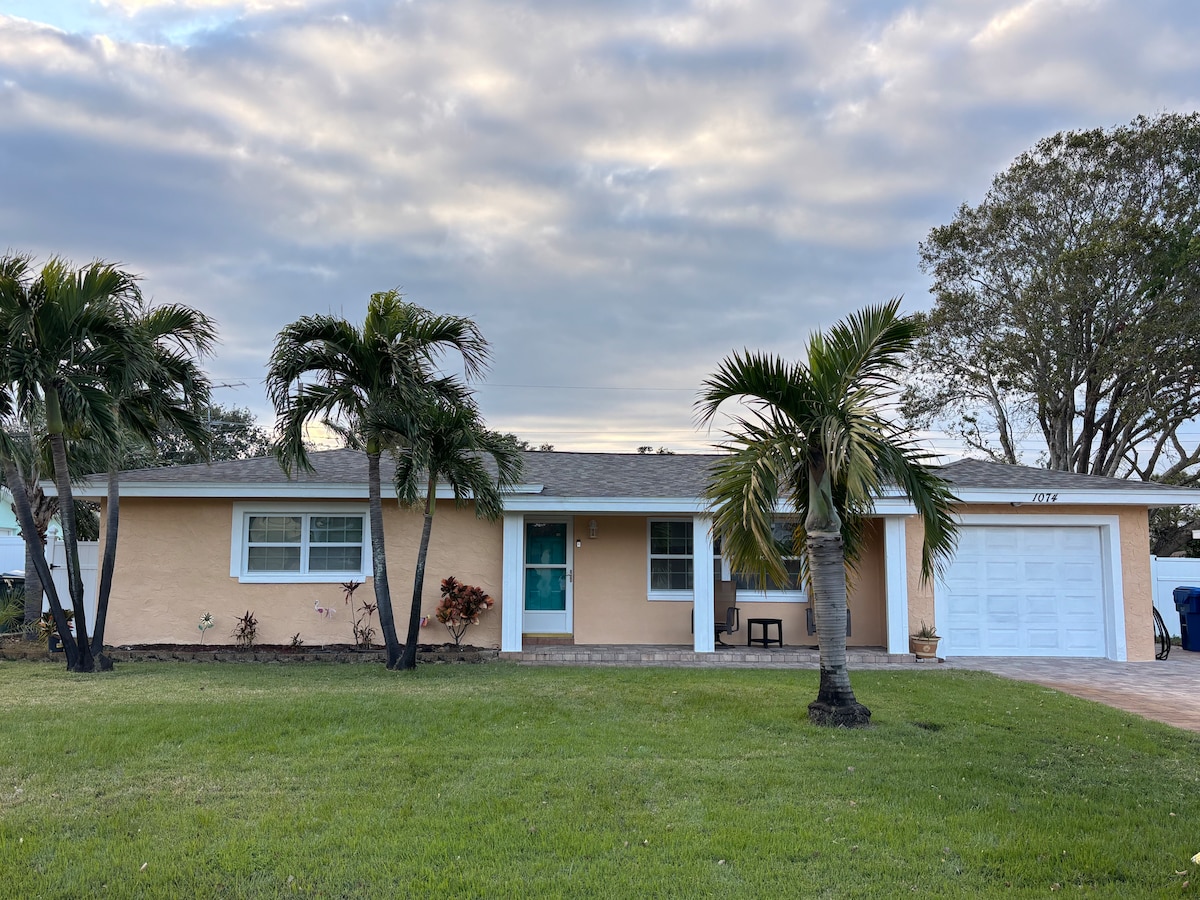 A single-story home is set amidst lush greenery, featuring a light-colored facade with a blue front door. Palm trees frame the entrance, while a manicured lawn extends before the house. A garage door is visible on the right side, adding to the inviting exterior.