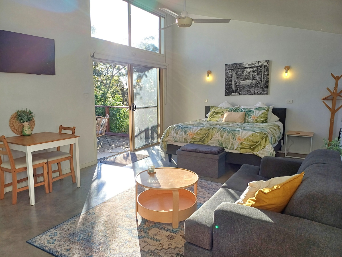 The open-plan suite features a spacious layout, showcasing a comfortable seating area with a grey sofa and a coffee table. A partially visible dining table is positioned nearby. Light streams through large glass doors that open to a private courtyard, enhancing the inviting atmosphere.