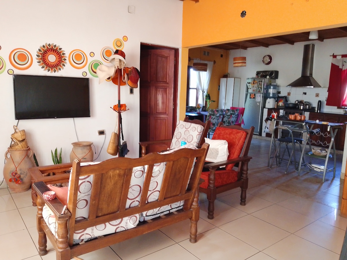 The living area features a comfortable seating arrangement with patterned cushions set against a bright orange wall. A flat-screen television is mounted on the wall, while an open kitchen area with modern appliances is visible in the background. Decorative elements contribute to the inviting atmosphere.