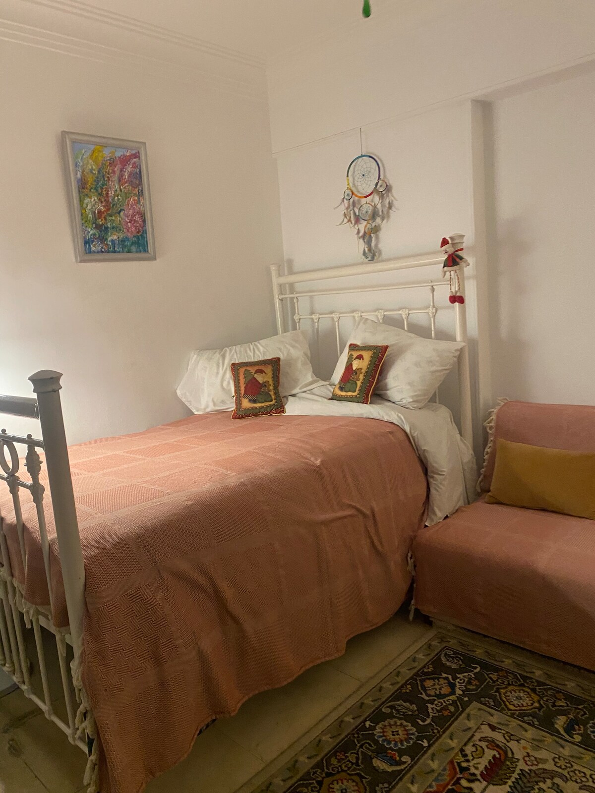 A private room features a small double bed dressed in crisp white linens, complemented by decorative pillows. A sofa bed is positioned nearby, with a colorful throw. Wall art adds a touch of personality, enhancing the clean, inviting atmosphere of the space.