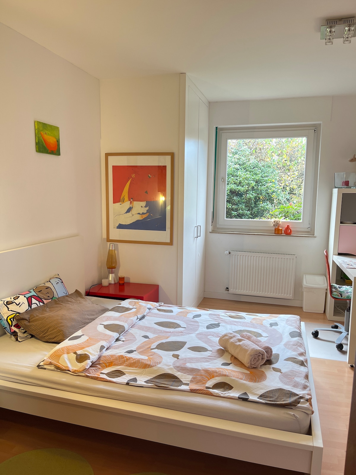 A cozy room is equipped with a spacious bed adorned with patterned linens. A window provides natural light and a view of greenery outside. A colorful piece of art hangs on the wall, complemented by a lamp and a small red furniture piece.