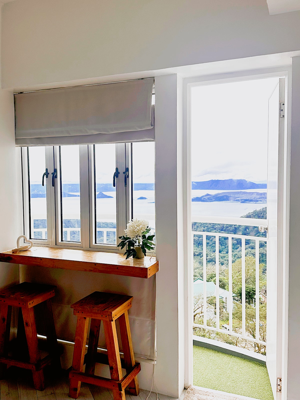 A window nook with two wooden stools is set against a wall, offering a view of Taal Lake and Volcano. Natural light illuminates the space, where a small vase with flowers adds a touch of freshness. A door leads to a balcony with scenic views.