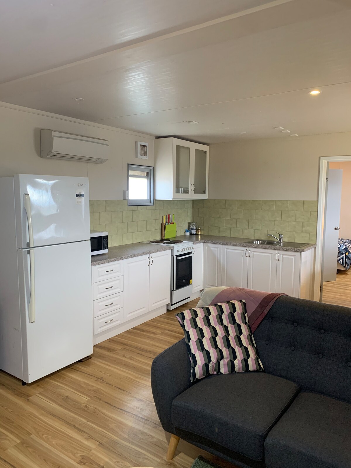 A modern kitchen area features white cabinetry and a light green tiled backsplash. Key appliances include a refrigerator, microwave, and oven. A cozy seating area is highlighted by a sofa with a patterned throw pillow, complemented by wooden flooring that adds warmth to the space.