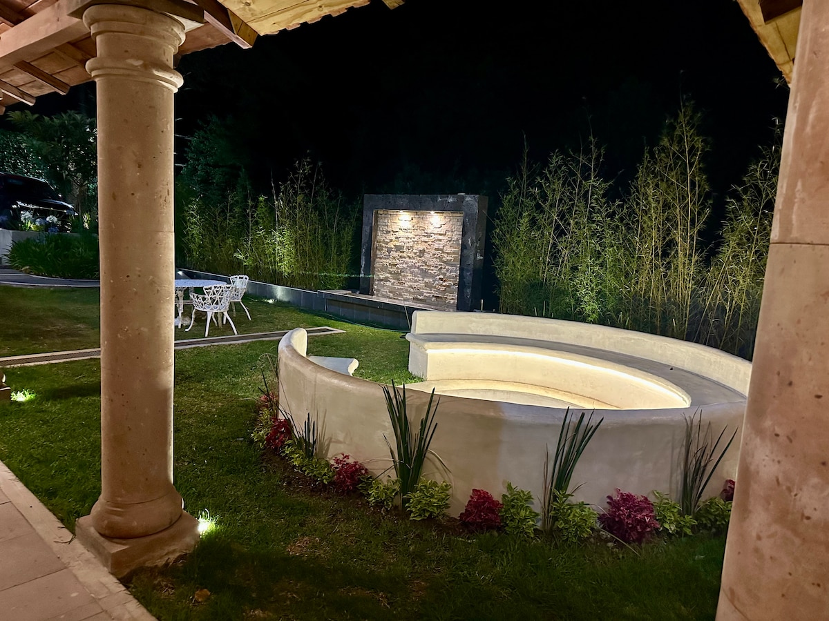 An outdoor seating area features a circular stone bench, softly illuminated for evening enjoyment. Lush greenery surrounds the space, complemented by a stone wall adorned with subtle lighting. A dining table with white chairs is positioned nearby on the manicured lawn.